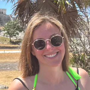 Smiling woman wearing sunglasses and a neon green swimsuit top, standing outdoors with palm trees and ancient ruins in the background on a sunny day.